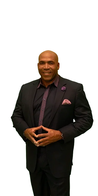 Bald, muscular Black man in a dark suit over a patterned shirt, smiling, with hands clasped in front.